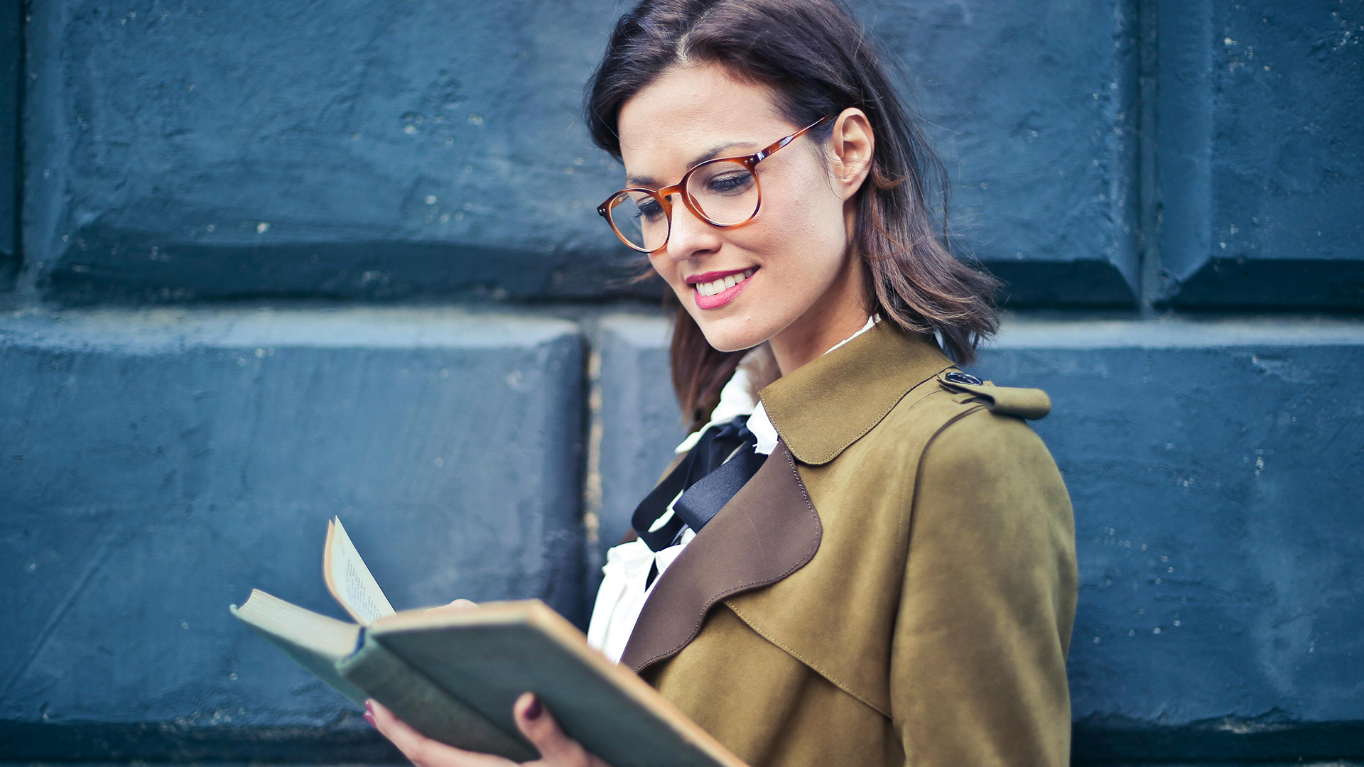 A woman wearing glasses, smiling while reading a book outdoors