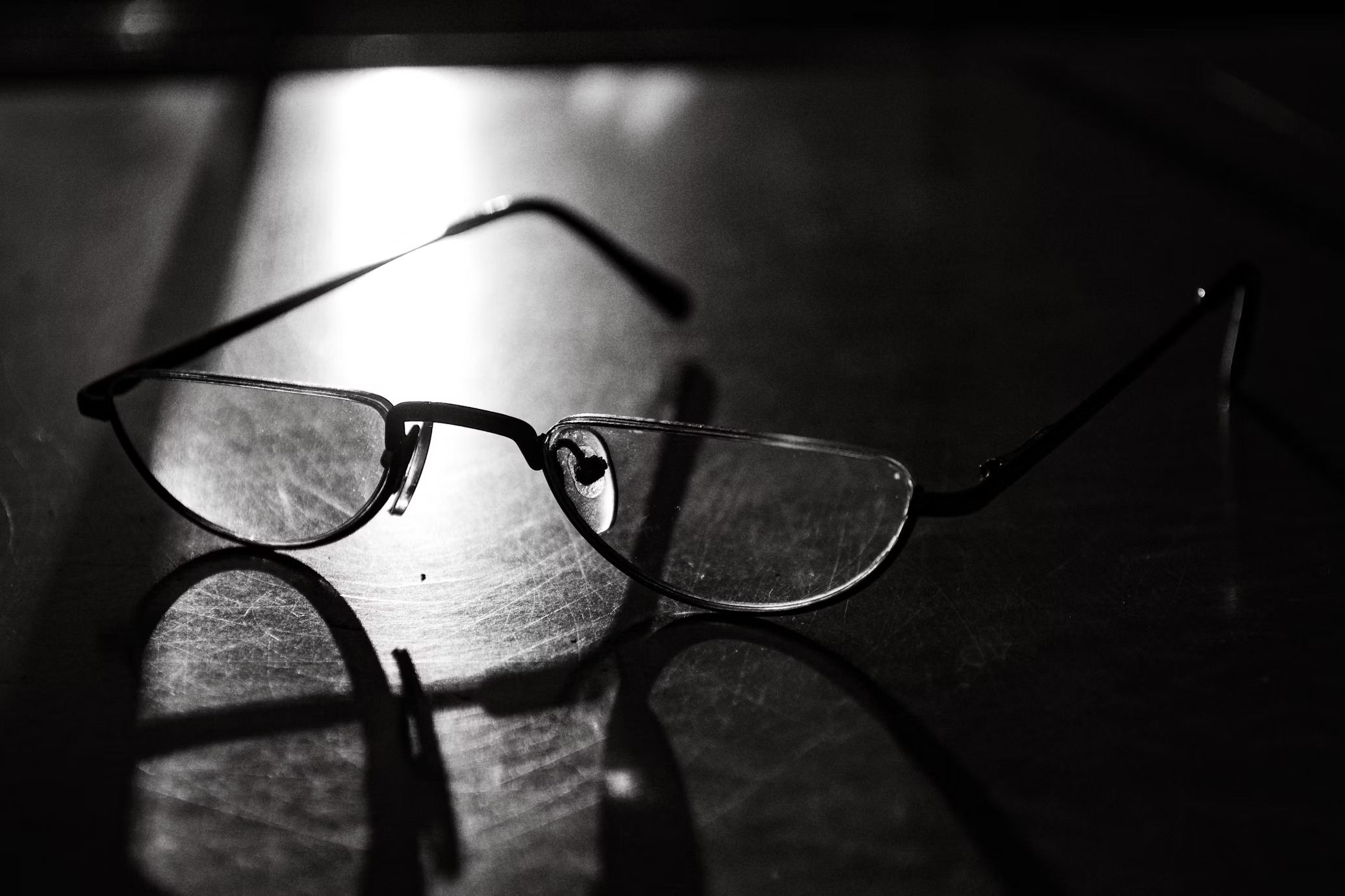 A pair of glasses resting on a reflective surface with shadows cast in the background